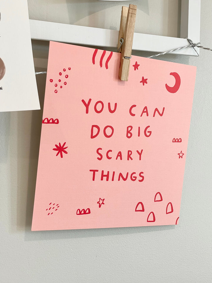 Pink poster with 'You can do big scary things' text and doodles, attached to a white wall with a clothespin.