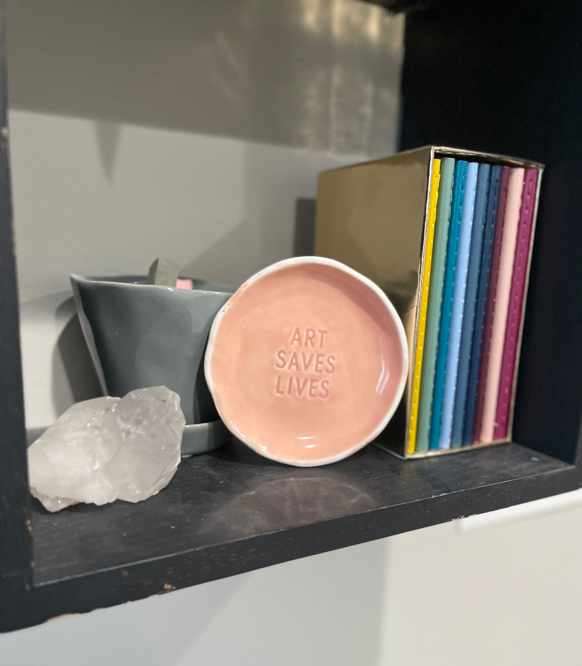 Shelf in artist studio showing a crystal, colorful journals and a pink dish that says Art Saves Lives.