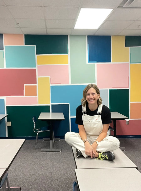 Artist Emily Frushour sitting in front of the bright and geometric mural she created for a classroom. 