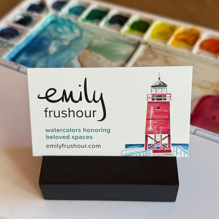 Business card for artist Emily Frushour, with a watercolor lighthouse and paints in the background.
