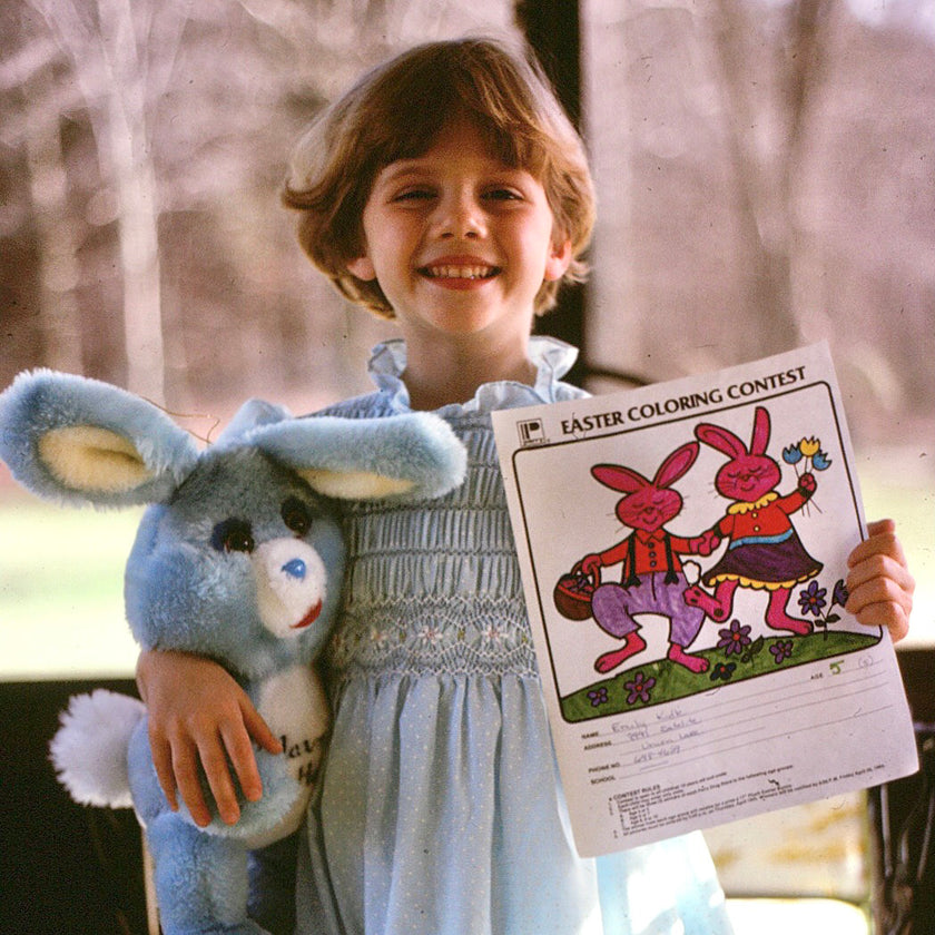Artist Emily Frushour as a young child, holding a drawing and a stuffed animal from a coloring contest.