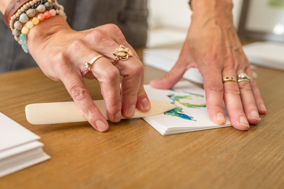 Artist Emily Frushour's hands folding notecards featuring a bright watercolor painting of the Great Lakes.