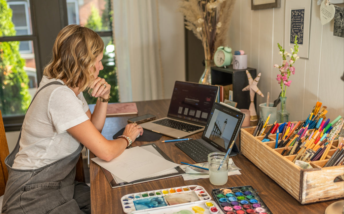 Artist Emily Frushour sitting at desk, working on computer with colorful art supplies and paints on the tabletop.