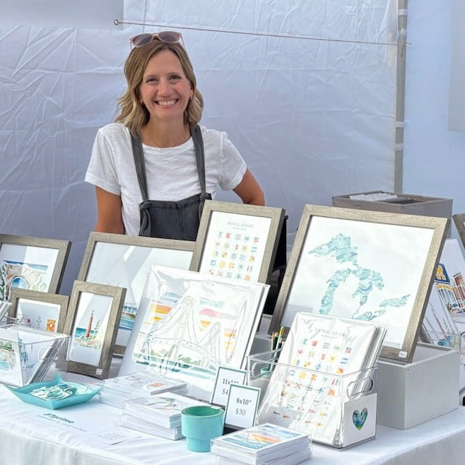 Artist Emily Frushour standing with her watercolor work in her booth at a summer art fair.