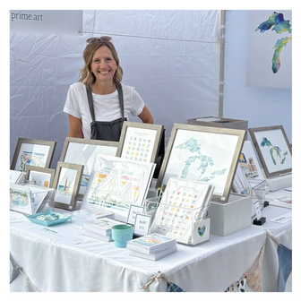 Artist Emily Frushour standing in a booth at the Grand Haven Coast Guard Market, selling her watercolor prints and notecards.