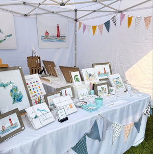 Artist Emily Frushour's booth display at a summer art market in Michigan, showing watercolor prints and notecards.