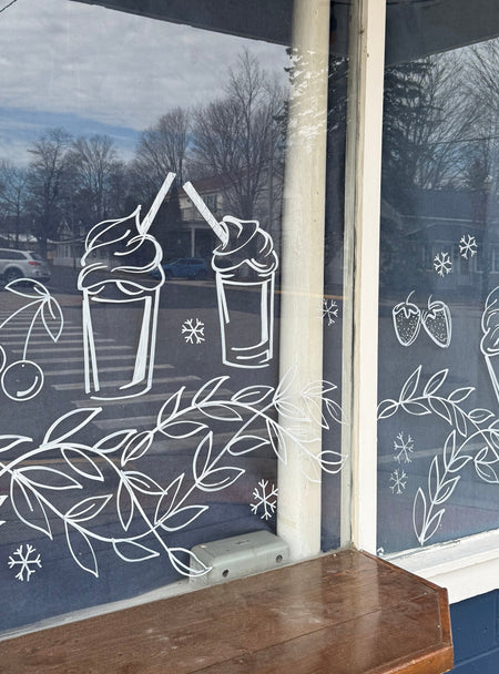 Delicate window art illustration at an ice cream shop showing snowflakes, milkshakes, fruit, and garland, created by artist Emily Frushour.