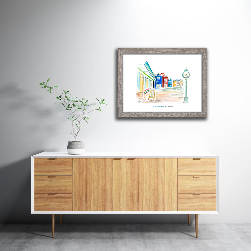 Bright and colorful watercolor painting of a street scene in Suttons Bay, Michigan, showing shops and a clock tower. Painting is hung in a gray frame in a living room.