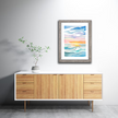 Framed colorful and bright watercolor painting of a lake at sunset in shades of blue, pink and yellow, hanging on a wall.