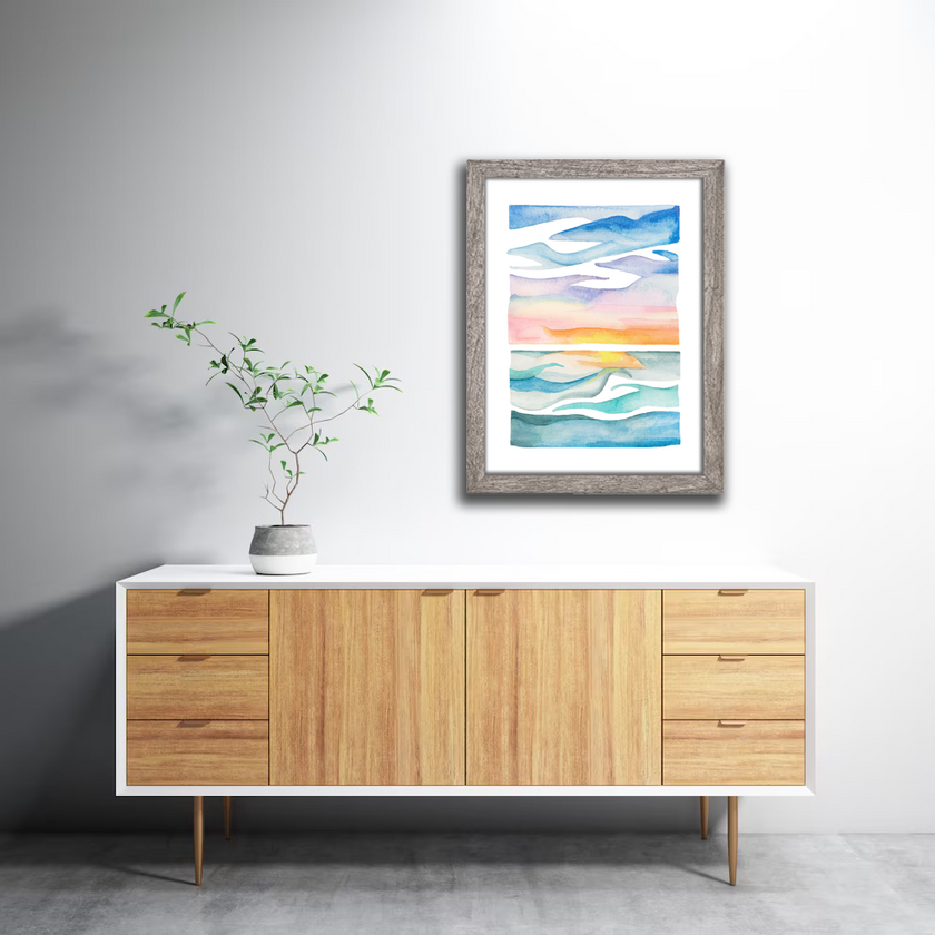Framed colorful and bright watercolor painting of a lake at sunset in shades of blue, pink and yellow, hanging on a wall.