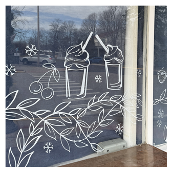 Delicate window art illustration at an ice cream shop showing snowflakes, milkshakes, fruit, and garland, created by artist Emily Frushour.