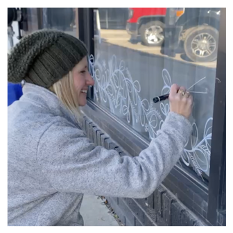 Artist Emily Frushour using chalk markers to create seasonal window art illustrations at a bakery.